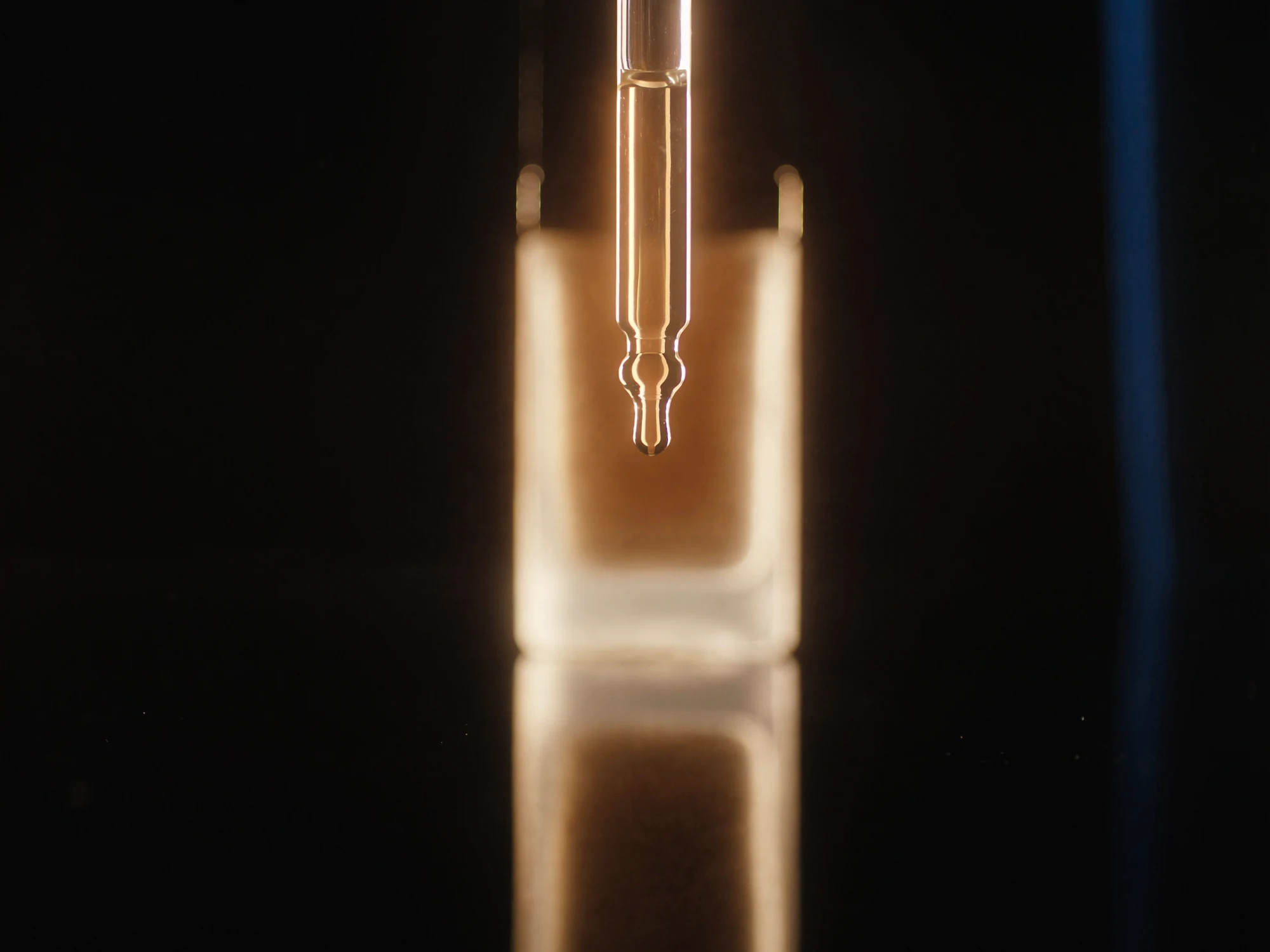 Goutte de sérum cosmétique ambré en suspension sous une pipette en verre — macro beauté de luxe par Fabien Bouadi